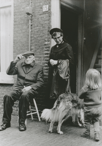 876925 Afbeelding van een Zuster Augustines naast een oudere man, die op een kruk zit bij de deur van zijn woning met ...